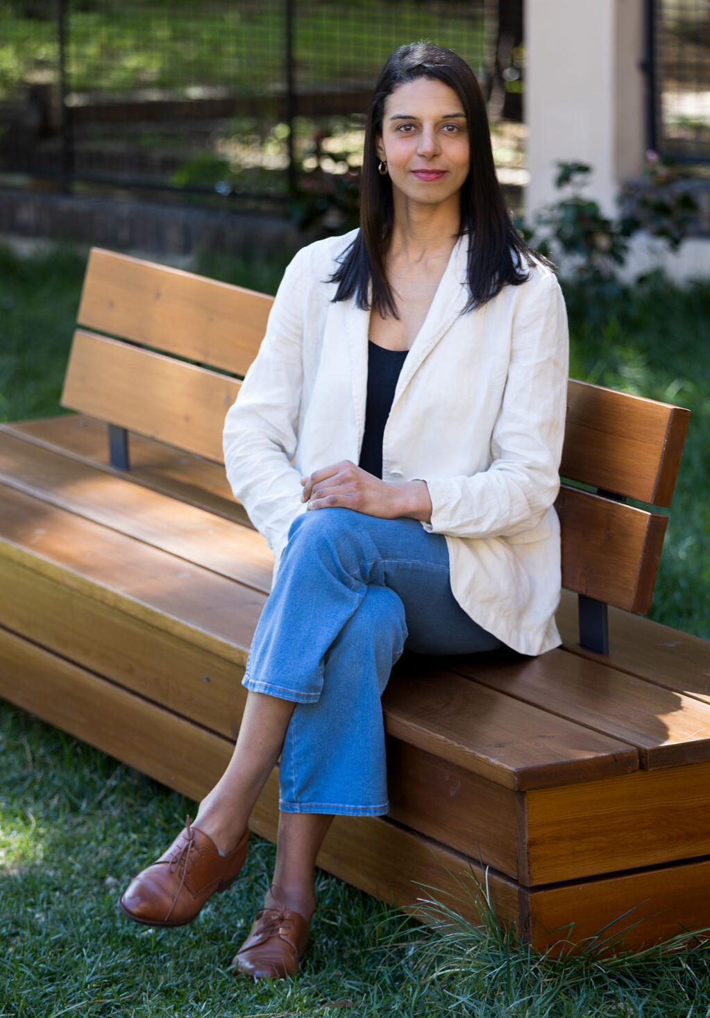 Nur Saeed sitting on a brown long chair, wearing a white jacket, blue jeans trouser, and a brown color shoe